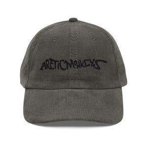 Puede incluir: Gorra de béisbol de pana verde oliva con el texto "ARCTIC MONKEYS" bordado en negro en la parte delantera. La gorra tiene una visera curva y una correa ajustable en la parte trasera.