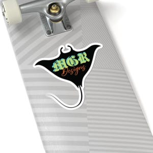 Puede incluir: Una pegatina con un diseño de manta raya negra con el texto "MGR Designs" en un degradado de verde, amarillo y naranja. La pegatina está sobre una patineta.