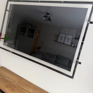 May include: A large, rectangular black framed mirror mounted on a white wall. The mirror reflects a room with furniture, including a grey sofa and a wooden cabinet. The frame is black and secured with visible screws.