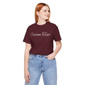 Pode incluir: Uma camiseta bordô com as palavras "Custom Text" em escrita branca. A camisa de manga curta é feita de um material macio e é combinada com jeans azuis. O estilo geral é casual e confortável.