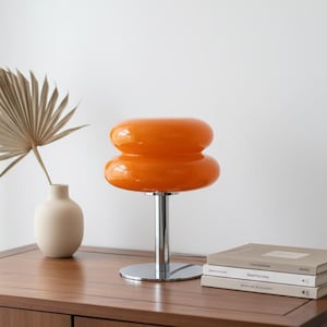 Peut inclure: Lampe de table orange de style rétro avec une base et une tige chromées. La lampe présente un design empilé et arrondi. Elle est posée sur une surface en bois, avec un vase et une plante séchée en arrière-plan.