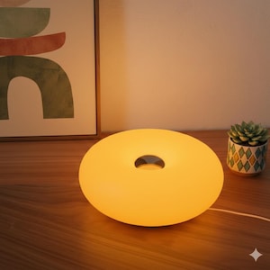 May include: A modern, donut-shaped table lamp with a warm yellow glow. The lamp has a circular design with a small, dark opening in the center. A small succulent plant in a patterned pot sits nearby.