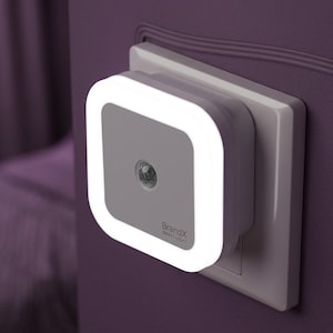 Puede incluir: Una luz nocturna cuadrada para enchufe con un borde brillante blanco y un panel central gris. El dispositivo cuenta con un sensor de movimiento circular en el centro y el texto BrandX SMART LIGHT impreso en la esquina inferior derecha.
