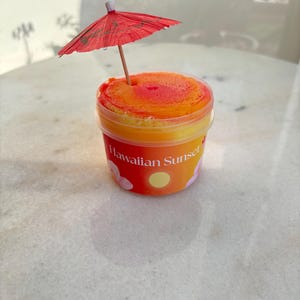 以下が含まれることがあります： オレンジと黄色のテクスチャーのあるスライムが入った透明なプラスチック容器で、上部には小さな赤い紙製のカクテル用傘が刺さっています。容器の前面のラベルには、白いフォントで「Hawaiian Sunset」という文字と、黄色い太陽とピンクの花のグラフィックが描かれています。
