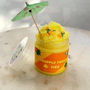 May include: A jar of yellow Pineapple Paradise Fizz with a green and orange label. The jar is topped with yellow foam and pineapple-shaped sprinkles. A small green umbrella with red floral accents is inserted into the foam. The lid is off to the side.