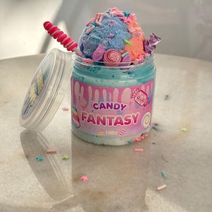 Peut inclure: Un pot de slime coloré avec le texte "CANDY FANTASY" sur le devant. Le slime est garni de slime bleu, rose et orange, de vermicelles et de bonbons. Une sucette rose et blanche est posée sur le côté.