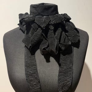 May include: A black pleated neck accessory featuring a high, structured collar and a voluminous, ruffled front section that cascades downward into two long, narrow, pleated fabric tails. The entire piece is made of a textured, accordion-pleated fabric.