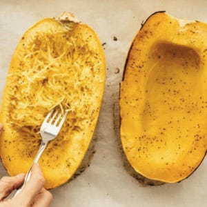 Puede incluir: Dos mitades de calabaza espagueti asada, con un tenedor desmenuzando los hilos de calabaza. La calabaza es de color amarillo-naranja con condimento de pimienta negra. La imagen está tomada desde arriba.