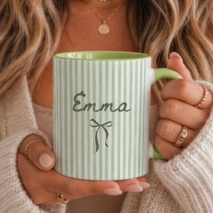 Peut inclure: Une tasse en céramique blanche à rayures verticales vert clair, avec un intérieur et une anse verts. La tasse porte le nom "Emma" en écriture cursive avec un motif de nœud. La tasse est tenue par une personne portant un pull crème.