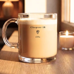 May include: A clear glass mug filled with a creamy drink, with the text "Chapter 18" above a floral illustration and the name "Ella." A lit candle is next to it on a wooden surface, creating a warm atmosphere. The mug has a large handle.