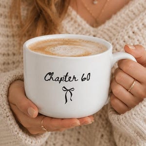 May include: A white ceramic mug filled with coffee, featuring the black printed text Chapter 60 and a small black bow graphic centered on the front. The mug has a rounded handle and a smooth finish, showcasing a clean and simple design.