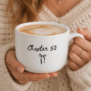 Peut inclure: Une tasse en céramique blanche remplie de café, avec le texte imprimé en noir "Chapter 50" au-dessus d'une petite icône de nœud noir. La tasse possède une anse arrondie et une finition lisse, tenue par deux mains contre un pull en maille crème.