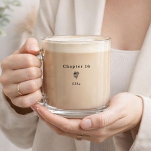 Peut inclure: Une tasse en verre transparent remplie d'une boisson crémeuse, peut-être du café ou du thé, avec une mousse sur le dessus. La tasse porte l'inscription "Chapter 16", une illustration florale et le nom "Ella".