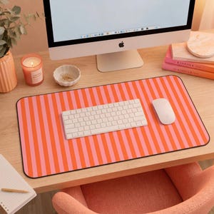 Op de afbeelding: Een rechthoekige bureaumat met een herhalend verticaal streeppatroon in afwisselende tinten feloranje en lichtroze. De mat ligt op een houten bureau onder een wit draadloos computertoetsenbord en een witte computermuis.