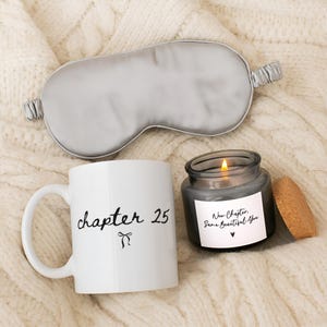 May include: A white ceramic mug with the black text "chapter 25" and a small bow icon, a grey satin sleep mask, and a dark glass candle with a lit wick and a label reading "New Chapter. Same Beautiful You" with a small heart icon.