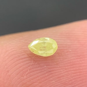 Pode incluir: Uma pedra preciosa em forma de pêra, facetada, amarelo claro. A gema está colocada sobre um fundo desfocado, com foco no corte e na cor da pedra. A pedra preciosa é de cor amarelo pálido.