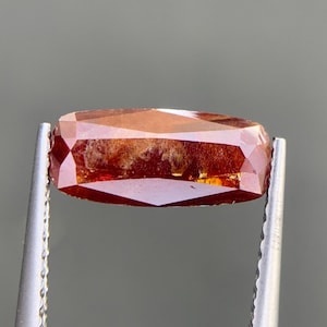 May include: A rectangular, faceted gemstone with a deep reddish-orange hue and visible internal inclusions. The stone is held by a pair of silver-toned metal tweezers, showcasing its elongated shape, polished surfaces, and translucent quality.