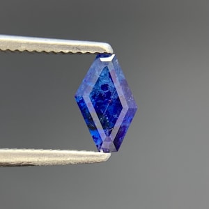 May include: A kite-shaped gemstone with a faceted surface, displaying a deep blue colour with internal mottled patterns and dark inclusions. The stone is held by metal tweezers, showcasing its geometric cut and vibrant, saturated blue hues.