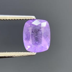 May include: A cushion-cut lavender gemstone with a faceted surface held by a pair of metal tweezers. The stone displays a translucent light purple hue with visible internal inclusions and sharp geometric facets reflecting light across its top.