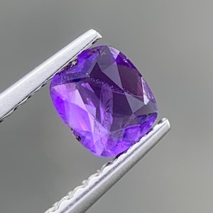 Könnte beinhalten: Ein kissenförmig geschliffener Amethyst-Edelstein mit leuchtend violetter Farbe und facettierter Oberfläche, gehalten von einer silberfarbenen Metallpinzette. Der Edelstein zeigt innere Klarheit und Lichtreflexionen auf seinen polierten Facetten.