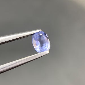May include: A loose oval-cut gemstone with a light periwinkle blue hue and faceted surface, held securely by a pair of silver-toned metal tweezers. The stone displays internal clarity and light reflection across its polished, symmetrical facets.