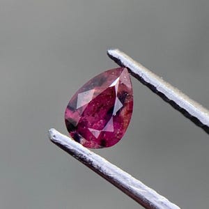 Puede incluir: Una piedra preciosa facetada en forma de pera con una paleta de colores magenta intenso y rojo oscuro. La piedra presenta inclusiones internas y está sujeta por unas pinzas de metal plateado, mostrando su calidad translúcida y corte.