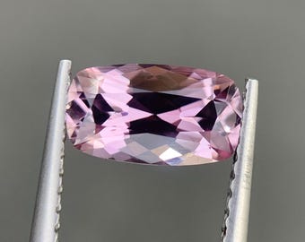 Peut inclure: Une pierre précieuse taille coussin aux teintes rose clair et lavande, dotée d'une surface à facettes qui reflète la lumière. La pierre est maintenue par une pince en métal argenté avec une prise dentelée, révélant sa clarté et sa forme.