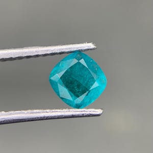 Puede incluir: Una gema facetada de color verde azulado, sujeta con pinzas plateadas. La gema tiene talla cojín, de forma cuadrada con esquinas redondeadas. Las facetas reflejan la luz, creando un efecto brillante. El fondo es gris neutro.