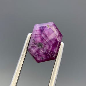 May include: A hexagonal, translucent magenta-colored gemstone with visible internal white striations and a dark inclusion near the center. The stone features a flat, polished surface with distinct geometric facets and sharp edges, displaying a deep pinkish-purple hue throughout its crystalline structure.