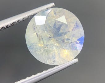 Peut inclure: Une pierre précieuse de pierre de lune ronde et facettée présentant un aspect blanc laiteux translucide avec des inclusions internes et de subtils reflets bleus et jaunes sur sa surface polie, tenue par une pince en métal argenté.