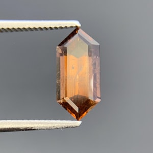 Könnte beinhalten: Ein facettierter Edelstein im Hexagonschliff mit einer tiefen bernsteinfarbenen und braunen Färbung. Der Stein weist scharfe, geometrische Kanten und eine durchscheinende Qualität auf, die innere Klarheit und Lichtreflexion auf der polierten Oberfläche zeigt.