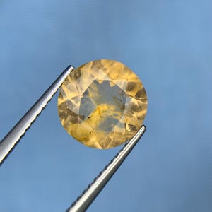 Puede incluir: Una piedra preciosa amarilla redonda y facetada con inclusiones internas visibles y patrones naturales de tonos dorados. La piedra translúcida está sujeta por unas pinzas de metal plateado, mostrando su superficie clara y multifacetada.