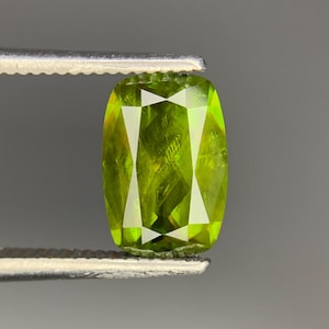 Könnte beinhalten: Ein kissenförmig geschliffener Peridot-Edelstein in leuchtendem Limettengrün mit facettierter Oberfläche, der sicher zwischen den Metallspitzen einer Pinzette gehalten wird. Der Stein zeigt innere Klarheit und Lichtreflexionen.