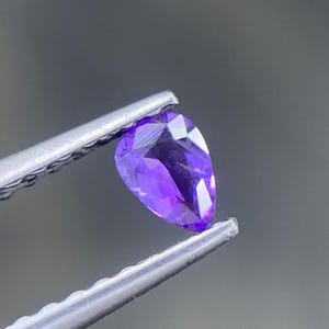 May include: A pear-shaped, faceted gemstone with a vibrant, deep purple color. The stone is held by a pair of silver-toned metal tweezers, showcasing its translucent quality and sharp, geometric cut facets that reflect light across its surface.