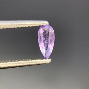 May include: A pear-shaped, faceted gemstone with a light purple hue. The stone features a pointed top and a rounded base, displaying internal clarity and light-reflecting facets throughout its surface, held securely by a metal tool.