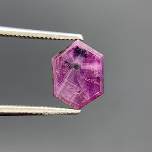 May include: A hexagonal cut gemstone with a vibrant magenta and deep pink hue. The stone features visible internal inclusions and natural veining patterns throughout its translucent surface, held securely by a pair of metal tweezers.