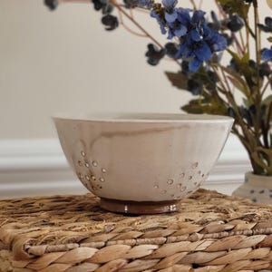 May include: A cream-colored ceramic bowl with a wide rim and a dark brown base. The bowl has small, evenly spaced holes around the sides. It sits on a woven wicker mat, with blue and yellow flowers in the background.