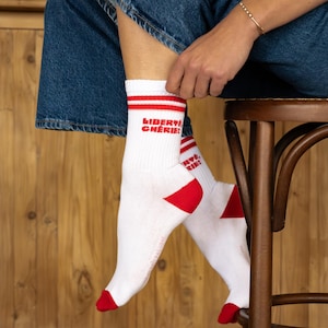 Peut inclure: Chaussettes blanches avec des détails rouges et le texte "LIBERTÉ CHÉRIE!" en rouge. Les chaussettes ont des rayures rouges en haut et des talons et orteils rouges. La personne porte un jean bleu et un bracelet doré.