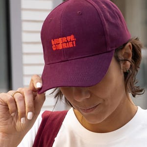 Könnte beinhalten: Burgunderfarbene Baseballkappe mit dem orangefarbenen Text "LIBERTÉ, CHÉRIE!". Die Kappe ist aus weichem Stoff gefertigt und hat einen gebogenen Schirm. Ein modisches Accessoire.