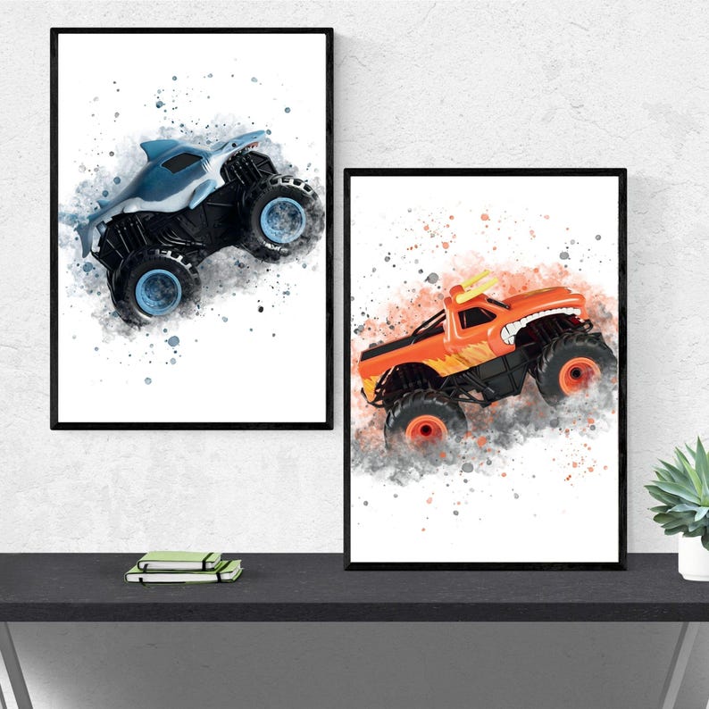 May include: Two framed watercolour prints of monster trucks. One shows a blue shark-themed truck, the other an orange truck with a toothy grin. Both are set against a white background with grey and blue splatters.