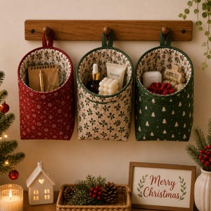 May include: Three fabric hanging storage pockets suspended from a wooden wall rack. The left pocket is red with white snowflakes, the center is cream with holly leaves, and the right is green with white Christmas trees, all holding various items.