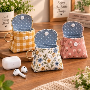 Può includere: Tre piccole pochette in tessuto con anelli in metallo dorato e chiusure a scatto bianche. Una pochette è a quadretti gialli, una è bianca con motivi floreali blu e gialli e una è rosa con stampe di margherite bianche.