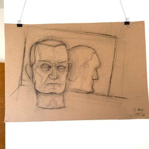 May include: A pencil sketch on brown paper shows a man's face and a profile. The man in the foreground wears glasses and has a serious expression. The artwork is held by binder clips, and the date 17.10.26 is visible.