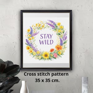 May include: A framed cross-stitch pattern with a floral wreath design. The wreath features yellow sunflowers, purple lavender, and orange and pink flowers. The words "STAY WILD" are in the center. The frame is black and the pattern is 35 x 35 cm.