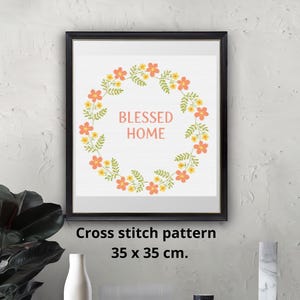 May include: Framed art print featuring a floral wreath with orange and yellow flowers and green leaves. The words "BLESSED HOME" are in the center. The frame is black. The cross stitch pattern is 35 x 35 cm.