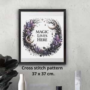 May include: Framed cross stitch pattern with a circular design featuring crystals, crescent moons, and the text "MAGIC LIVES HERE." The artwork is 37 x 37 cm. The frame is black, and the design includes purple, black, and white elements.