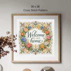 Op de afbeelding: Een ingelijst kruissteekpatroon met de tekst "Welcome to our home". Het ontwerp toont een bloemenkrans met blauwe, oranje, roze en gele bloemen. De lijst is lichtbruin van kleur. Het patroon meet 96.5 x 96.5 cm.