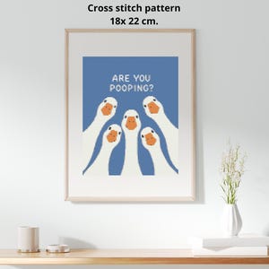 Può includere: Uno schema a punto croce di 18x22 cm raffigurante cinque oche bianche con becchi arancioni su sfondo blu. Il testo ARE YOU POOPING? è scritto in lettere maiuscole bianche sopra le oche. Il disegno è presentato all'interno di una cornice in legno chiaro.