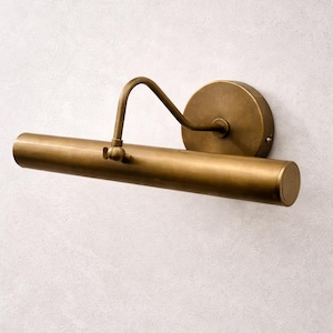 May include: A brass-toned picture light fixture. The fixture has a cylindrical light bar, a curved arm, and a circular wall mount. The finish is a warm, metallic color, and the design is simple and elegant.