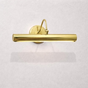 May include: A brass picture light fixture with a cylindrical light bar and a curved arm. The fixture is mounted on a round base and casts a shadow on the white wall. The light bar is approximately 12 inches long.